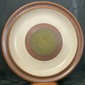 以下が含まれることがあります： 茶色の縁取りと、茶色、緑、白の濃淡で描かれた同心円模様が施された、大きな丸い陶器の皿です。
