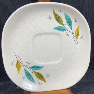 Peut inclure: Une soucoupe en céramique blanche avec un centre carré et une finition mouchetée. La soucoupe présente un motif stylisé de feuilles vertes, bleues et jaunes avec des tiges noires et des accents d'étoiles noires.