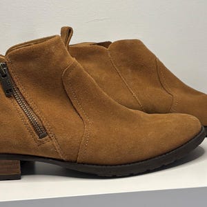 Puede incluir: Un par de botines de ante marrón con cremallera lateral y tacón pequeño. Las botas tienen una lengüeta en la parte trasera y la marca "UGG" es visible en el lateral.