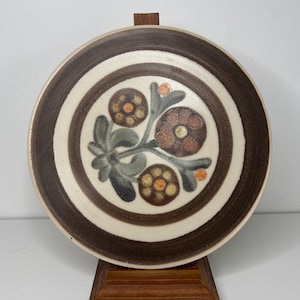 Puede incluir: Plato redondo de cerámica con diseño floral. El plato presenta anillos concéntricos de color marrón y crema. El centro del plato tiene un estampado floral en tonos marrón, naranja y verde. El plato se exhibe sobre un soporte de madera.