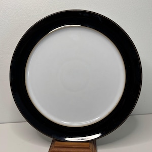 May include: A circular, white plate with a broad black border and a thin gold accent. The plate is displayed on a wooden stand. This elegant plate is suitable for serving meals. The plate's diameter is approximately 25 cm.