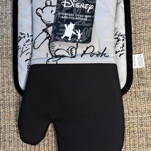 May include: Oversized oven mitt and pot holder set featuring Winnie the Pooh. The mitt is black, with a textured surface, and the pot holder is light grey with a black border. The set includes a Disney tag.