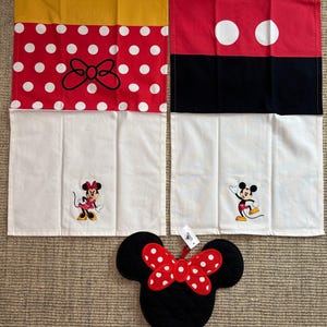 May include: Four Disney-themed kitchen textiles. Two red and white polka dot and yellow pieces, and two red, black, and white pieces. A black Mickey Mouse-shaped oven mitt with a red polka dot bow is also included. The items are on a neutral surface.
