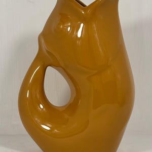 May include: A glossy mustard-yellow ceramic pitcher featuring a stylized fish-shaped silhouette. The handle is integrated into the body, forming a circular opening. The top rim is shaped like an open fish mouth, creating a distinct spout for pouring.