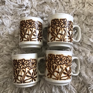 Può includere: Set di quattro tazze in ceramica bianca con un motivo floreale marrone. Le tazze hanno una forma semplice e arrotondata e un piccolo manico.
