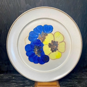 May include: A white ceramic plate featuring a blue and yellow floral design. The flowers are stylised and have a brown centre.