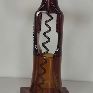 May include: A vintage brown and amber corkscrew with a bottle-shaped design. The corkscrew has a black metal worm and a rectangular base. The handle is a rectangular shape. The design is reminiscent of a wine bottle.