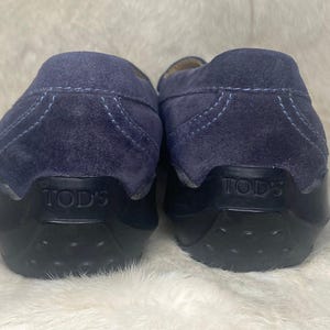 Puede incluir: Un par de mocasines de ante azul marino con suelas de goma negras. El nombre de la marca "TOD'S" est&aacute; grabado en el tal&oacute;n de cada zapato. Los zapatos se exhiben sobre una superficie blanca texturizada.