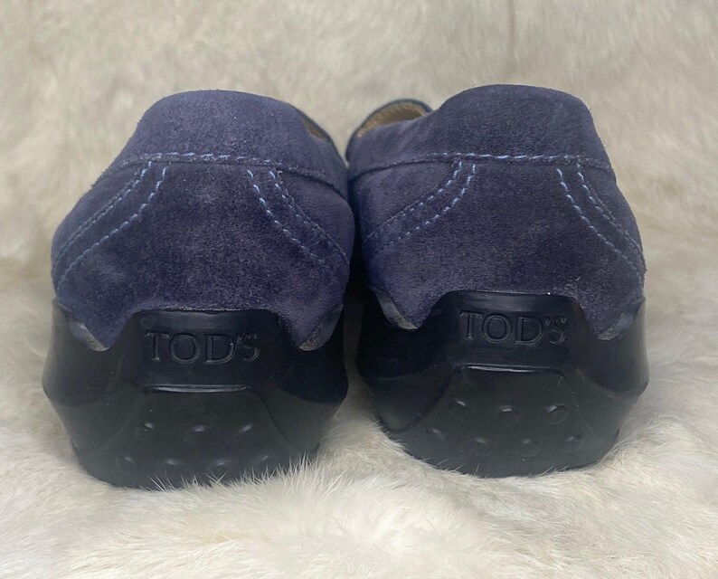 Puede incluir: Un par de mocasines de ante azul marino con suelas de goma negras. El nombre de la marca "TOD'S" est&aacute; grabado en el tal&oacute;n de cada zapato. Los zapatos se exhiben sobre una superficie blanca texturizada.