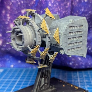 May include: A detailed model of a futuristic engine or spaceship component. The gray and gold-colored piece is mounted on a black base, set against a blue grid and a galaxy-themed backdrop. The model features intricate mechanical details.