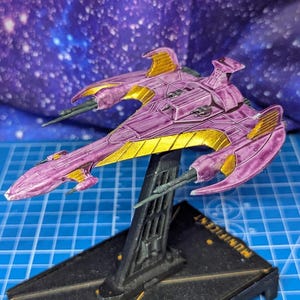May include: A purple and gold spaceship model on a black stand. The model has a sleek design with wing-like structures and is set against a starry background. The base has text on it.