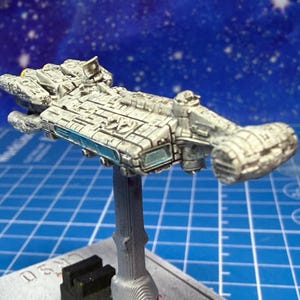 May include: A detailed model spaceship, painted in grey and off-white, mounted on a silver stand. The spaceship features intricate detailing, including panels, windows, and various protrusions. The background features a blue surface with a grid pattern and a starry sky.