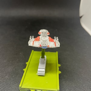 May include: A white and red spaceship model with a blue base. The spaceship has a rectangular body with a pointed nose and wings. The model is on a green base.