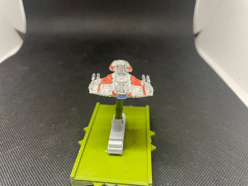 May include: A white and red spaceship model with a blue base. The spaceship has a rectangular body with a pointed nose and wings. The model is on a green base.