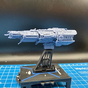 May include: A light blue model of a spaceship, mounted on a black stand. The spaceship has detailed features, including multiple sections and protrusions. The stand is on a blue cutting mat.