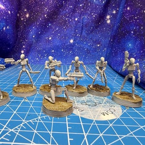 May include: A collection of gray miniature robot figures in various action poses, each holding a weapon. The figures stand on circular bases with textured, brown surfaces. The background features a blue surface with a starry pattern.