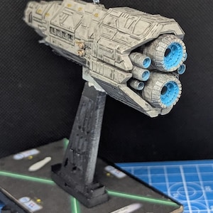 May include: A white and gray spaceship model with blue engine details. The model is mounted on a black stand and is displayed on a game board with a card that reads "Dreadnought Class, Vacuum Warp".