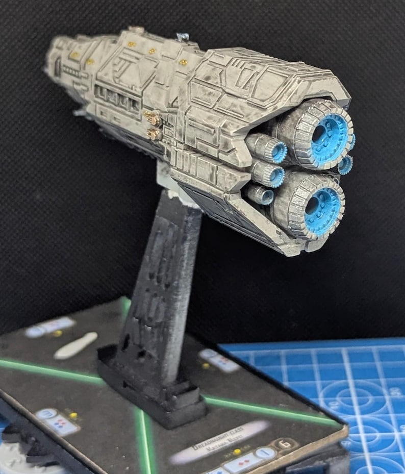 May include: A white and gray spaceship model with blue engine details. The model is mounted on a black stand and is displayed on a game board with a card that reads "Dreadnought Class, Vacuum Warp".
