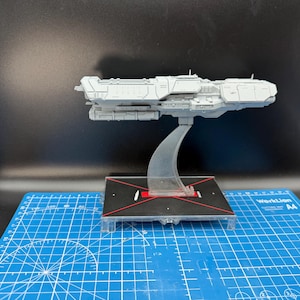 May include: A gray model spaceship, mounted on a clear stand, with a black and red base. The spaceship has detailed features and is likely a collectible or display item. The base sits on a blue cutting mat.
