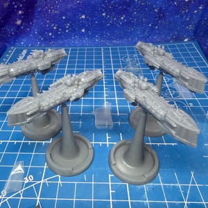 Könnte beinhalten: Vier graue Miniatur-Raumschiffmodelle auf Ständern, angeordnet auf einer blauen Rasteroberfläche. Die Modelle weisen detaillierte Designs auf und sind wahrscheinlich für ein Tabletop-Spiel oder Hobby gedacht. Ein klares Kunststoffteil ist ebenfalls vorhanden.