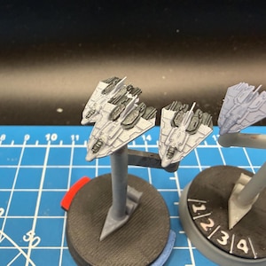 May include: Miniature grey spaceship models on stands, with detailed features. The models are on circular bases, some with red and blue accents. The background is a blue cutting mat with white grid lines and numbers.