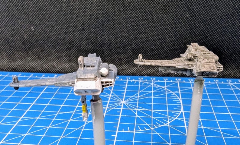 Op de afbeelding: Twee miniatuur-ruimteschepen geverfd in een grijs en wit kleurenschema. De ruimteschepen zijn gemonteerd op standaards en klaar om te worden tentoongesteld.