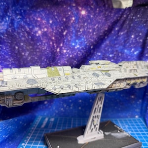 Könnte beinhalten: Ein detailliertes Modell eines Raumschiffs, hauptsächlich weiß mit grauen und grünen Akzenten, auf einem Ständer montiert. Das Modell weist komplizierte Verkleidungen und Designelemente auf, die auf ein Science-Fiction-Thema hindeuten. Der Hintergrund ist eine Sternengalaxie.