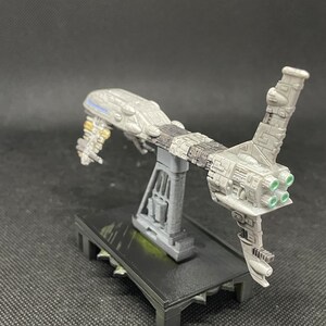 May include: A gray and white spaceship model with a stand. The spaceship has a long, thin body with a large, round cockpit. The spaceship has four green exhaust ports on the back. The stand is black and has a textured surface.