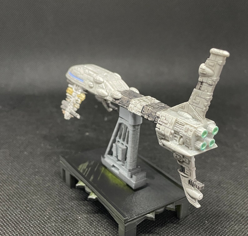 May include: A gray and white spaceship model with a stand. The spaceship has a long, thin body with a large, round cockpit. The spaceship has four green exhaust ports on the back. The stand is black and has a textured surface.