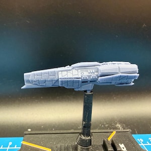 May include: A light blue model spaceship with intricate detailing, mounted on a black stand. The spaceship features a textured surface and various geometric shapes, suggesting a futuristic design. The model is set against a dark background.