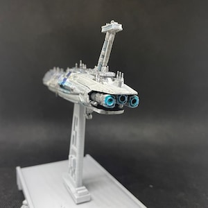 Puede incluir: Un modelo detallado de una nave espacial blanca y gris con puertos de escape azules. La nave espacial est&aacute; montada en un soporte gris.
