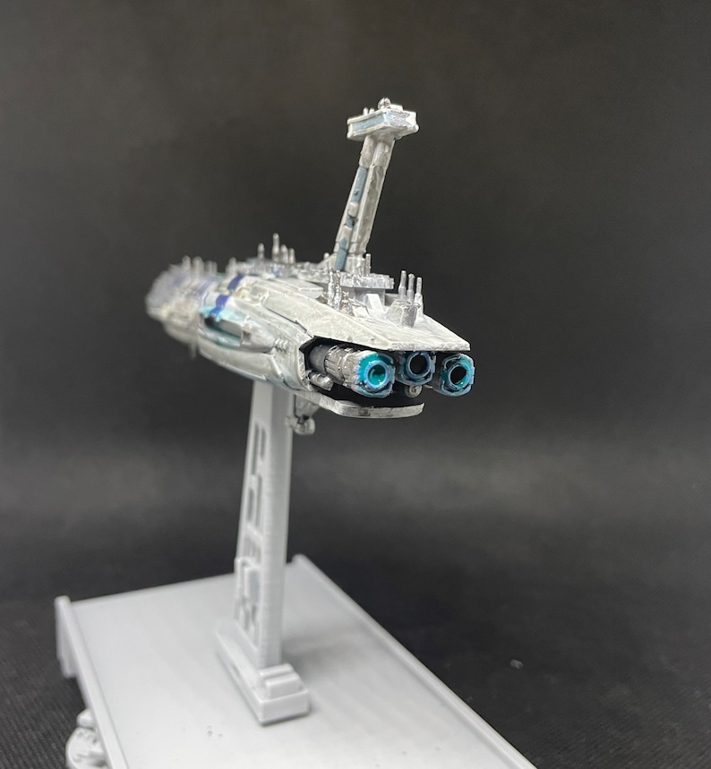 Puede incluir: Un modelo detallado de una nave espacial blanca y gris con puertos de escape azules. La nave espacial est&aacute; montada en un soporte gris.