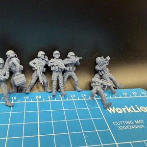 May include: A collection of gray miniature soldier figurines in various action poses. The figures are equipped with helmets, tactical vests, and rifles. The figures are on a blue cutting mat with the text "WorkLion CUTTING MAT 320X240mm".