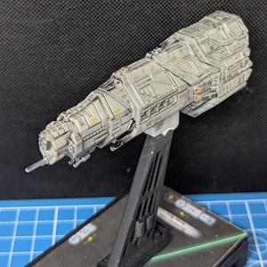 May include: A gray and white spaceship model with gold accents, mounted on a black stand. The spaceship has a long, cylindrical body with a pointed nose and a series of small windows.