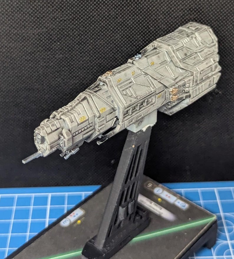 May include: A gray and white spaceship model with gold accents, mounted on a black stand. The spaceship has a long, cylindrical body with a pointed nose and a series of small windows.