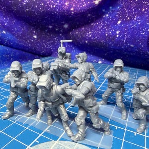 May include: A group of gray miniature figures, possibly soldiers or space troopers, are arranged on a blue grid surface. They are wearing helmets and tactical gear, some holding weapons. The background features a starry, purple fabric.