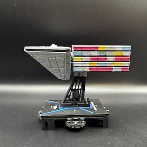 May include: A detailed model of a spaceship with a gray triangular front section and a colorful rectangular back section. The model is mounted on a black base with a support structure. The base has the word "REBEL" printed on it.