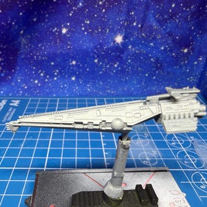 Puede incluir: Modelo gris claro de una nave espacial, con un cuerpo largo y triangular y componentes detallados del motor. El modelo está montado sobre un soporte, sobre un fondo azul estrellado. El modelo mide aproximadamente 25 cm de largo.