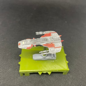 May include: A gray and red spaceship model on a green base. The spaceship has a detailed design with multiple windows and a pointed nose.