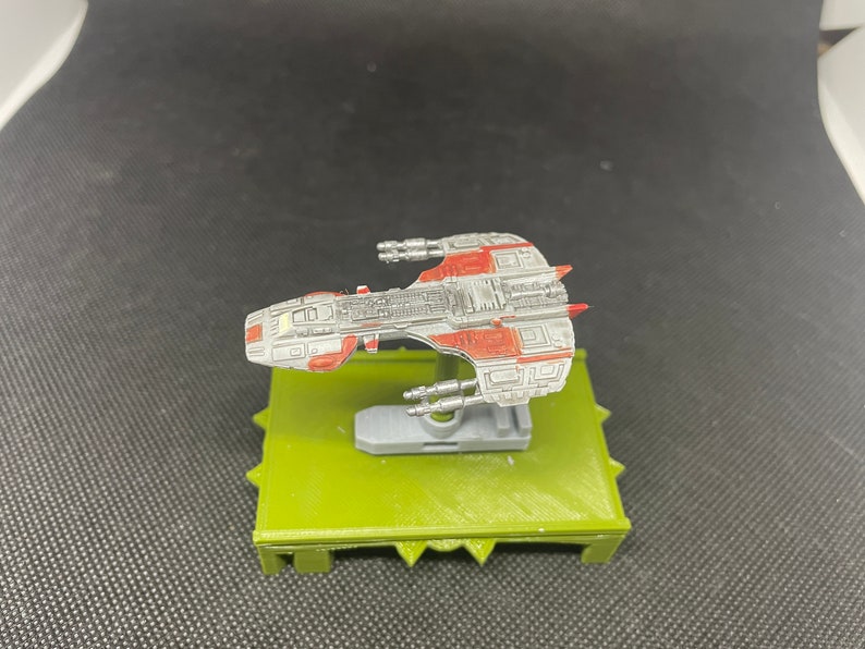 May include: A gray and red spaceship model on a green base. The spaceship has a detailed design with multiple windows and a pointed nose.