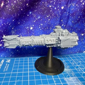 Könnte beinhalten: Graues, detailliertes Modell eines Raumschiffs, montiert auf einem schwarzen Ständer. Das Raumschiff weist komplizierte Designs auf und ist vor einem blauen Hintergrund mit Sternenmuster platziert. Das Modell ist wahrscheinlich ein Sammlerstück oder ein Spielobjekt.