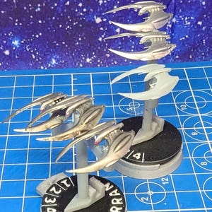 May include: Three silver painted miniature spaceships on black bases with white numbers. The bases are on a blue gridded surface. The spaceships are in a formation with the front ship facing forward and the two rear ships facing slightly to the side. The ships have a sharp, angular design with a pointed nose and wings.