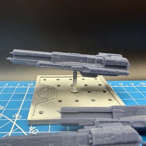 以下が含まれることがあります： グレーの3Dプリントされた宇宙船の模型で、スタンドに取り付けられています。宇宙船には、複数のセクションや窓と思われるものなど、詳細な特徴があります。スタンドは正方形で、円形のロゴが付いています。背景は青いカッティングマットです。