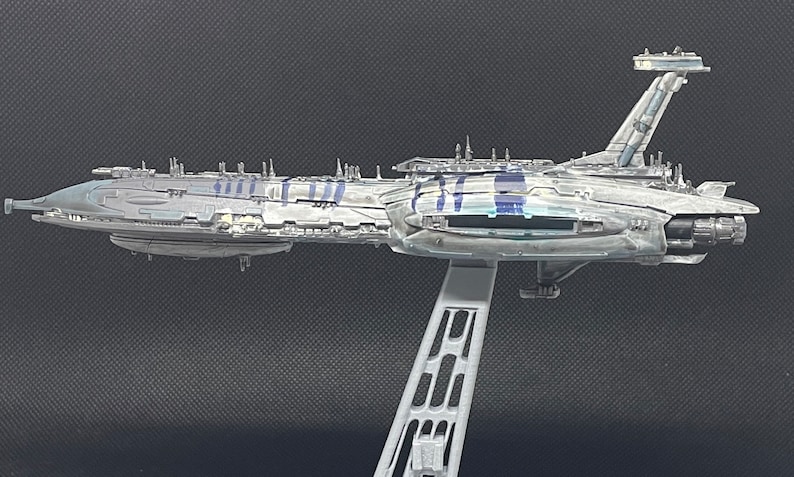 Puede incluir: Un modelo detallado de una gran nave espacial gris con un cuerpo largo y delgado y una aleta de cola vertical alta. El modelo est&aacute; montado en un soporte y parece estar hecho de pl&aacute;stico.