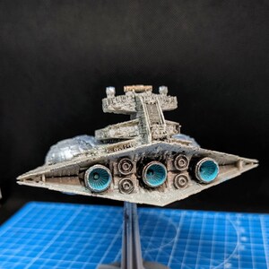 May include: A detailed model of a Star Destroyer spaceship, painted in a weathered silver and gray color scheme. The model is displayed on a black and gray stand, with the underside of the ship visible, showcasing the engine exhaust ports.