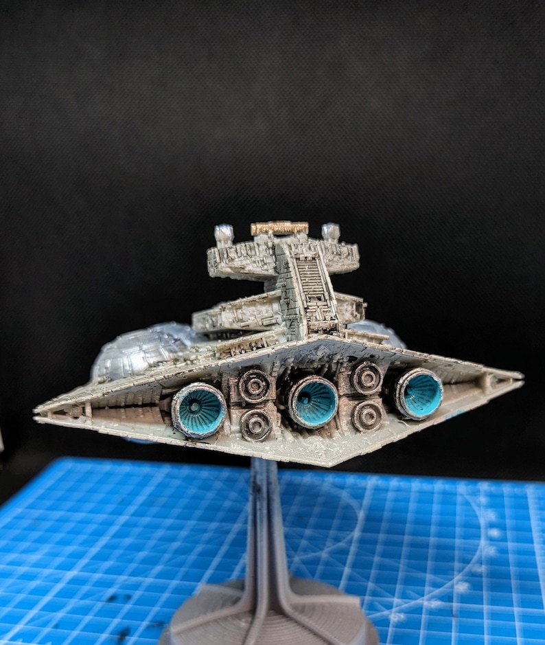May include: A detailed model of a Star Destroyer spaceship, painted in a weathered silver and gray color scheme. The model is displayed on a black and gray stand, with the underside of the ship visible, showcasing the engine exhaust ports.