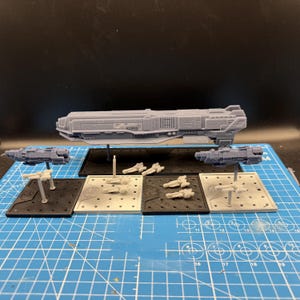 以下が含まれることがあります： グレーの3Dプリント製ミニチュア宇宙船のコレクション。黒いスタンドに乗った大型船1隻、スタンドに乗った中型船2隻、およびスタンドなしの小型船数隻が、グリッド線付きの青いカッティングマットの上に並べられています。