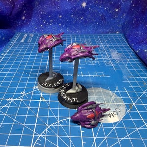 Puede incluir: Tres modelos de naves espaciales moradas en miniatura con detalles rojos, exhibidos sobre una superficie de cuadrícula azul. Dos están montados en soportes con bases numeradas, y uno descansa sobre la superficie. El fondo presenta una tela estrellada.