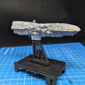 May include: A white and gray spaceship model on a black display stand. The spaceship has a detailed design with yellow accents. The stand has a rectangular base and a tall, open-frame support.