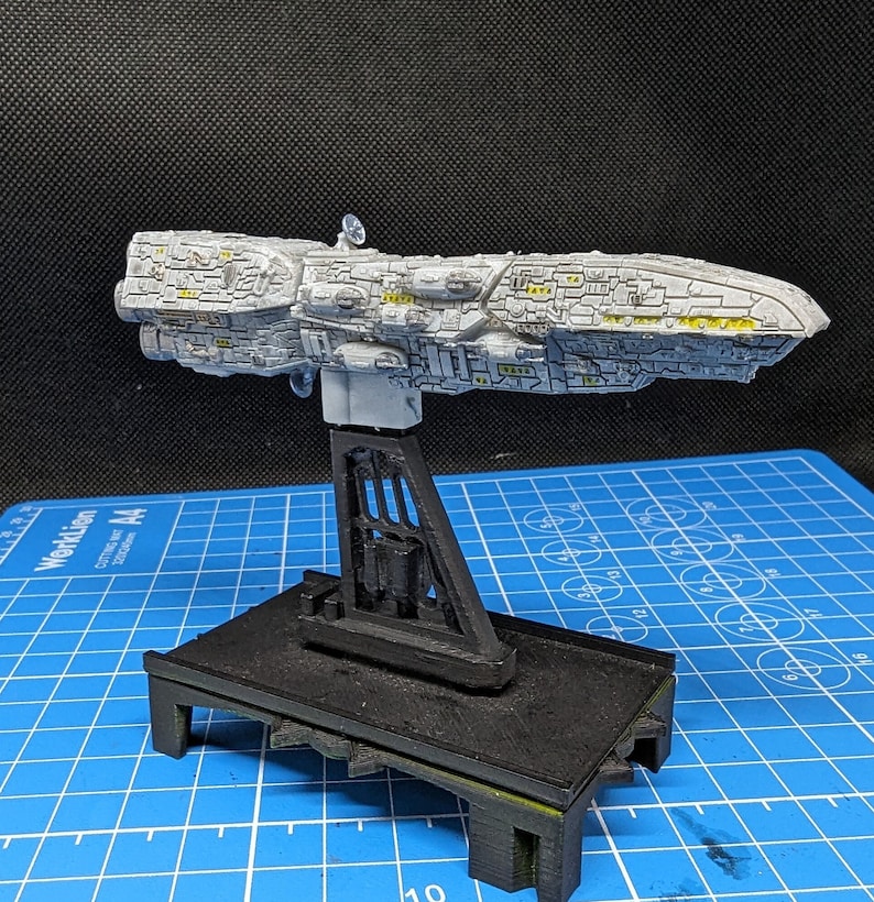 May include: A white and gray spaceship model on a black display stand. The spaceship has a detailed design with yellow accents. The stand has a rectangular base and a tall, open-frame support.